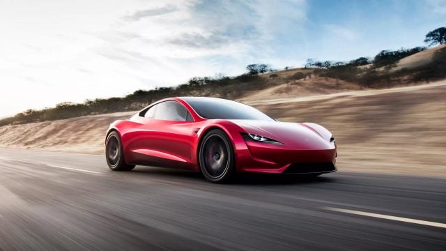 Tesla Roadster - czerwony samochód sportowy jedzie z dużą prędkością po asfaltowej drodze, z rozmytym tłem pokazującym suchy krajobraz i niebo.