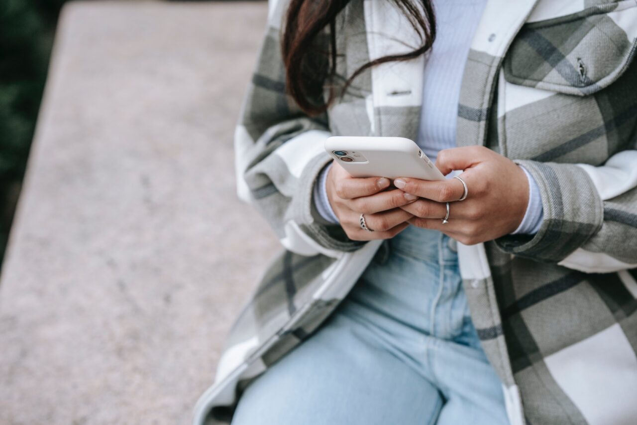 Zdjęcie do artykułu o tym, jak zwiększyć bezpieczeństwo w sieci - osoba siedzi na ławce i trzyma smartfona w rękach; widoczne są ręce, część kurtki w kratę, jeansy.