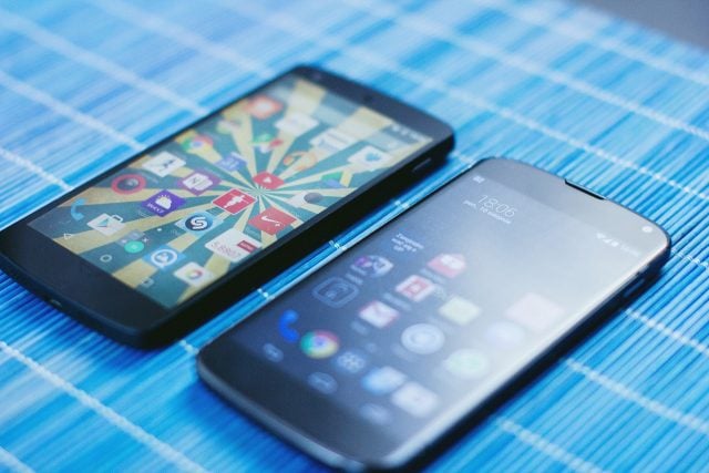 Dwa smartfony leżące na niebieskiej, w paski powierzchni. Jeden wyświetla ekran główny z ikonami aplikacji, a drugi zablokowany ekran z zegarem.