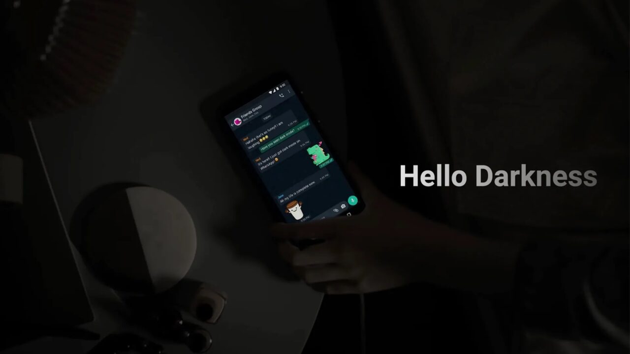 ciemny motyw na messengerze. Osoba trzymająca smartfon z włączoną aplikacją do komunikacji w ciemnym pomieszczeniu, na ekranie widoczne są czaty, a w prawym dolnym rogu napis "Hello Darkness".