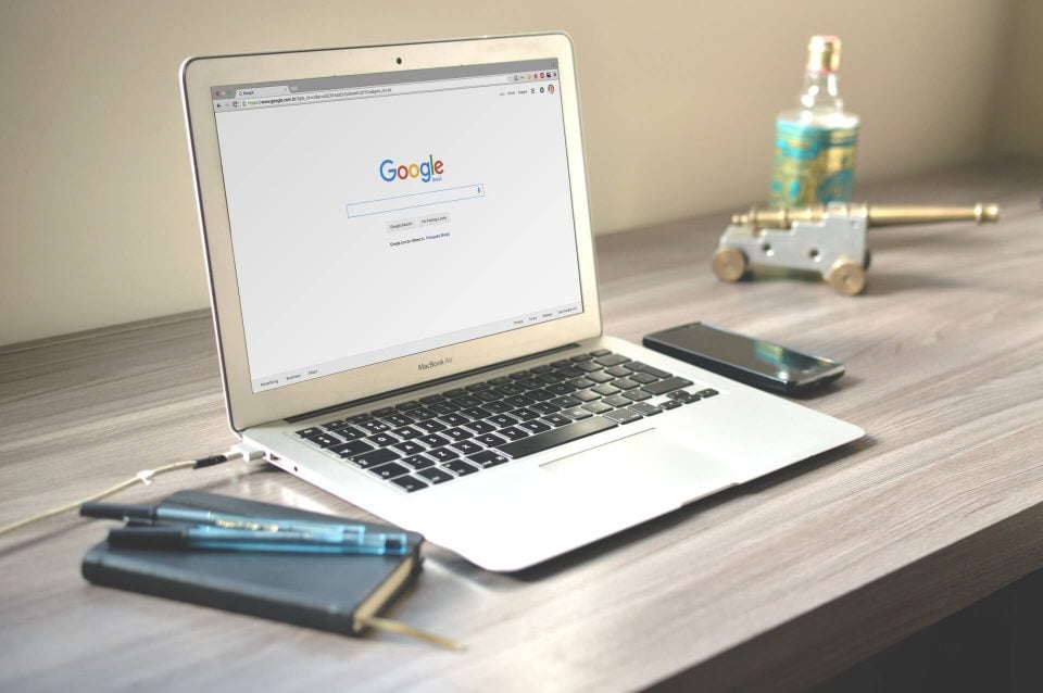 Laptop MacBook otwarty na stronie głównej Google na biurku, obok długopisy, smartfon i dekoracyjna butelka z płynem.