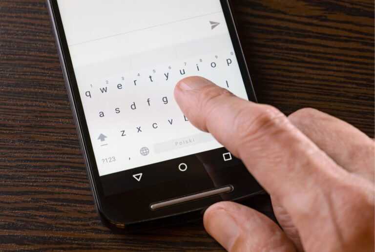 Dłoń osoby dotykającej ekran smartfona z widoczną na nim klawiaturą ekranową z napisem "polski" w prawym dolnym rogu.