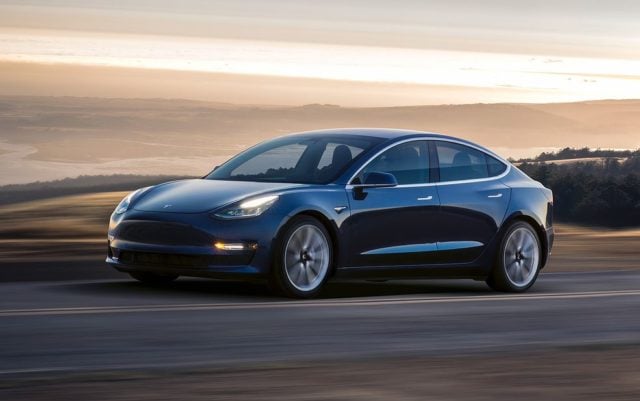 Niebieski samochód elektryczny marki Tesla Model 3 jedzie po drodze przy zachodzie słońca.
