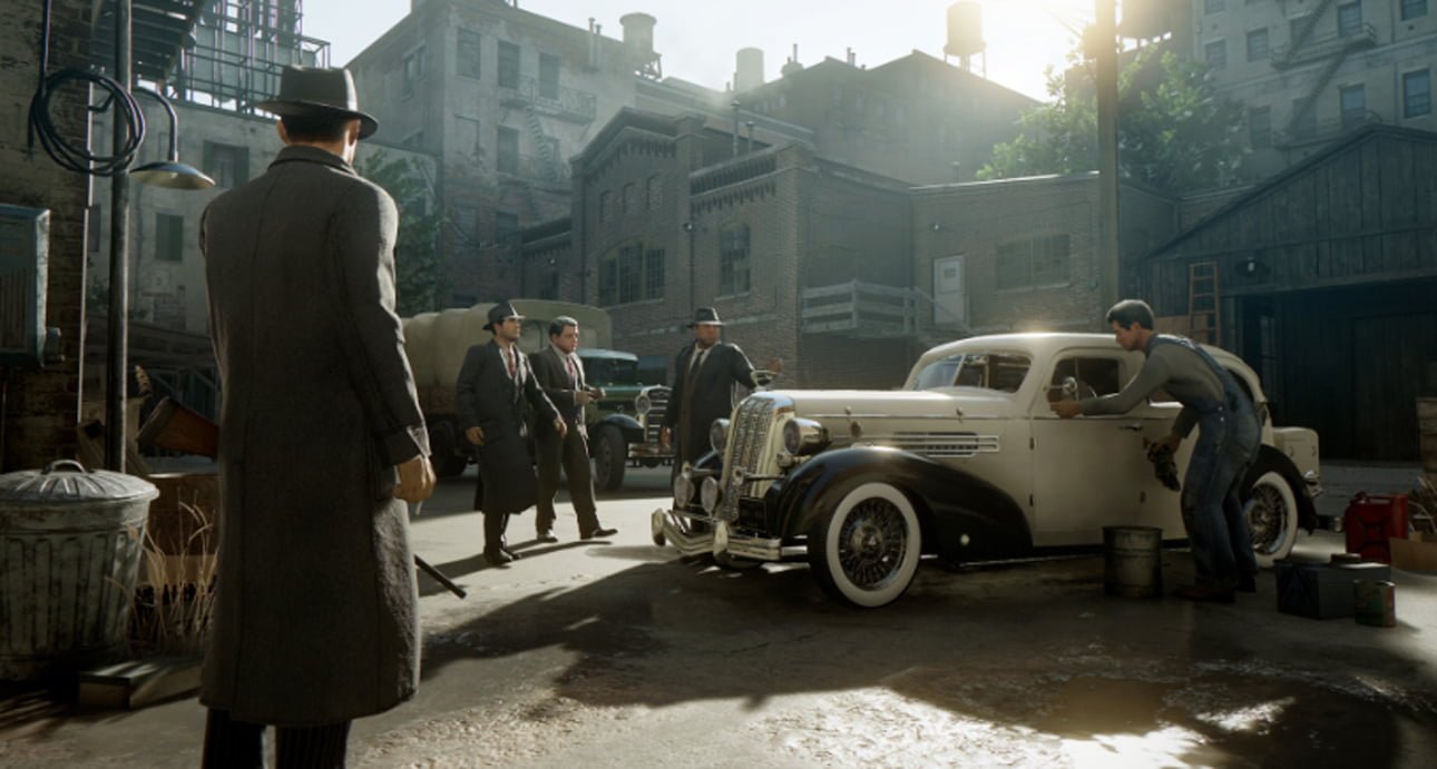 Rozgrywka z Mafia Definitive Edition. Scena uliczna z lat 30. XX wieku z mężczyznami w garniturach i kapeluszach podchodzącymi do zaparkowanego samochodu, podczas gdy mechanik pracuje przy innym pojeździe.