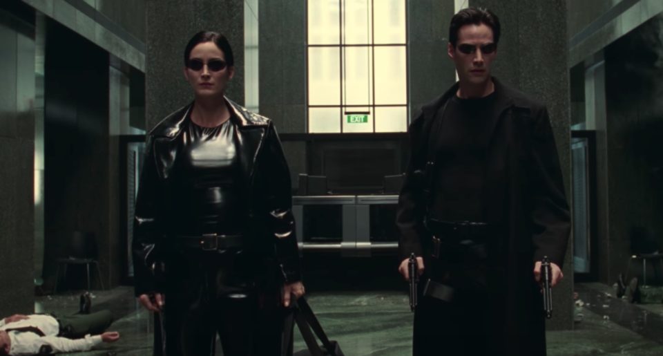 O czym opowie Matrix 5? Kobieta i mężczyzna w czarnych skórzanych płaszczach i okularach przeciwsłonecznych stoją w półmrocznym, nowoczesnym korytarzu, za nimi widoczny znak wyjście, na podłodze leży bezruchoma osoba.