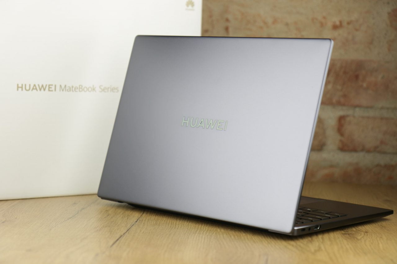 Laptop marki Huawei z serii MateBook ustawiony na drewnianym blacie z rozmytym tłem pokazującym pudełko z logo producenta.