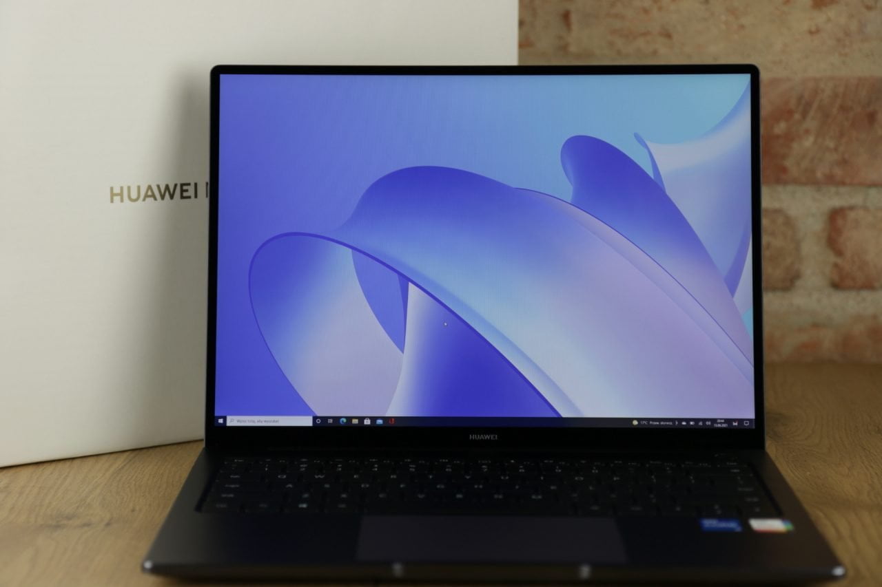 Laptop Huawei na stole z włączonym ekranem, na którym wyświetlony jest graficzny motyw w odcieniach niebieskiego, a za laptopem widoczne jest opakowanie z logo marki.