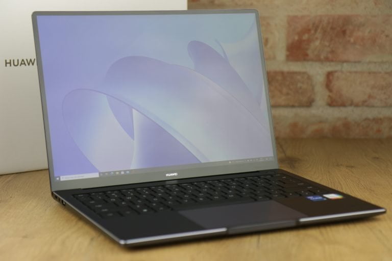 Laptop marki Huawei otwarty na drewnianym biurku z widocznym ekranem wyświetlającym grafikę i logo producenta pod ekranem; w tle ceglana ściana.