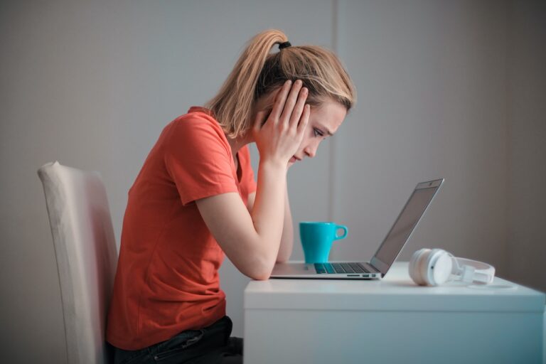 Kobieta w czerwonej koszulce siedzi przed laptopem z wyrazem zmartwienia na twarzy, trzymając głowę dłonią; na biurku obok niej niebieski kubek i białe słuchawki.