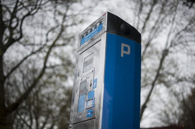 Niebieski automat do opłacania parkowania z napisem "PARKOMAT" na tle rozmytych drzew.