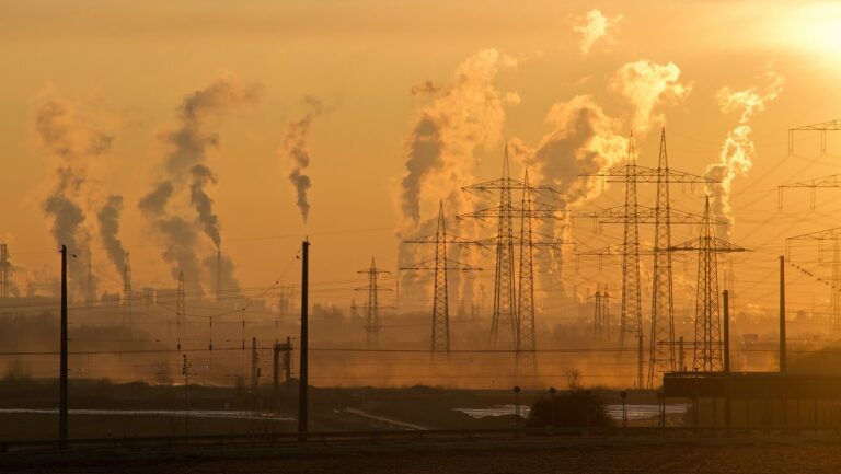 Zachód słońca nad przemysłowym krajobrazem z licznymi kominami fabrycznymi emitującymi dym oraz liniami przesyłowymi w tle. Strefa czystego transportu ma ukrucić takie widoki