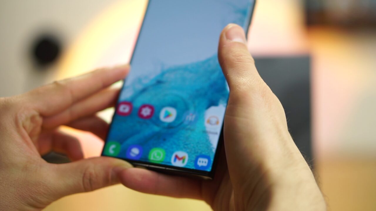 Dłonie trzymające smartfon Samsung Galaxy S22 Ultra 5G z kolorym, aktywnym ekranem startowym w pomieszczeniu.