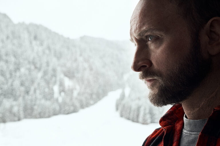 Mężczyzna w czerwonej koszuli w kratę patrzy przez okno na zaśnieżone góry.