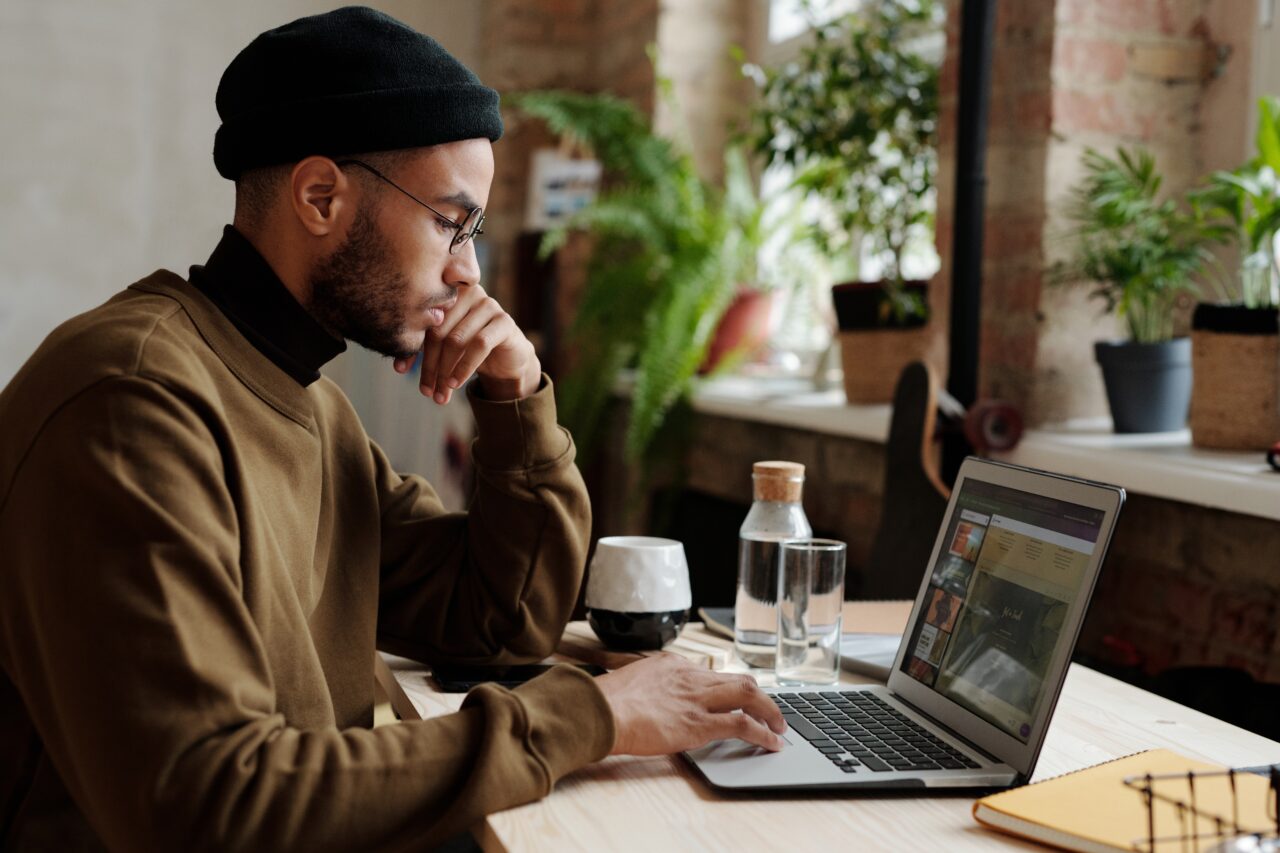 Mężczyzna w okularach i czarnej czapce pracujący na laptopie w przestronnym wnętrzu z roślinami w tle.