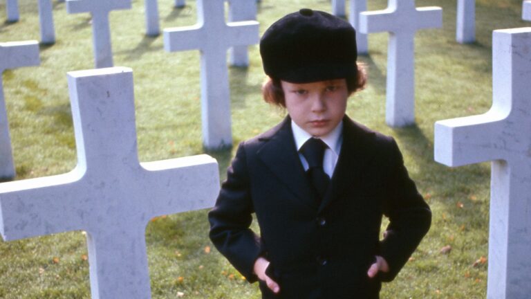 Kadr z filmu Omen. Dziecko w eleganckim ubraniu stoi przed białymi krzyżami na cmentarzu wojskowym.