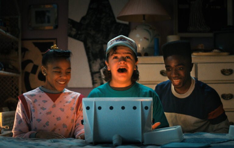 Trójka uśmiechniętych dzieci siedzi przed niebieskim komputerem w pokojowym otoczeniu przy słabym oświetleniu.