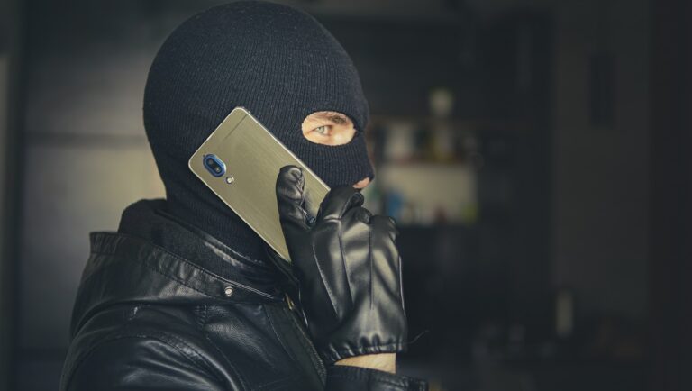 Osoba w kominiarce i czarnych skórzanych rękawiczkach przystawia srebrny smartfon do ucha.