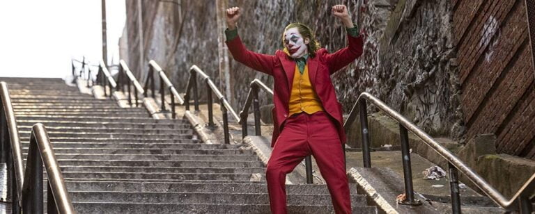 aktor grający Jokera w kostiumie klauna tańczy radośnie na schodach na zewnątrz.