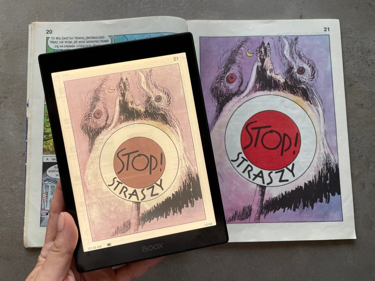 Osoba trzyma otwartą książkę z ilustracjami przedstawiającymi stylizowane potwory z napisami "STOP! STRASZY". Jedna strona jest wyświetlana na elektronicznym czytniku, a druga znajduje się w tradycyjnej książce.