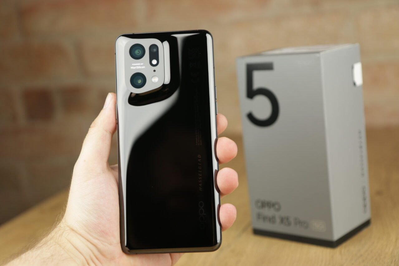 Czarna tylna klapka smartfona z zaawansowanym modułem aparatu trzymanego w dłoni, z rozmytym opakowaniem w tle.