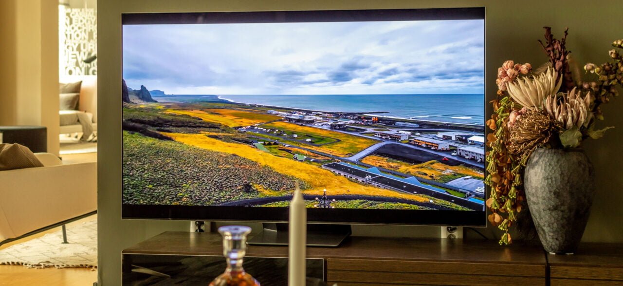 Telewizor samsung S95B powieszony na ścianie w pokoju. Na ekranie widać krajobraz przedstawiający niebo, wodę oraz tereny zielone. Obok telewizora stoi wazon z kwiatami