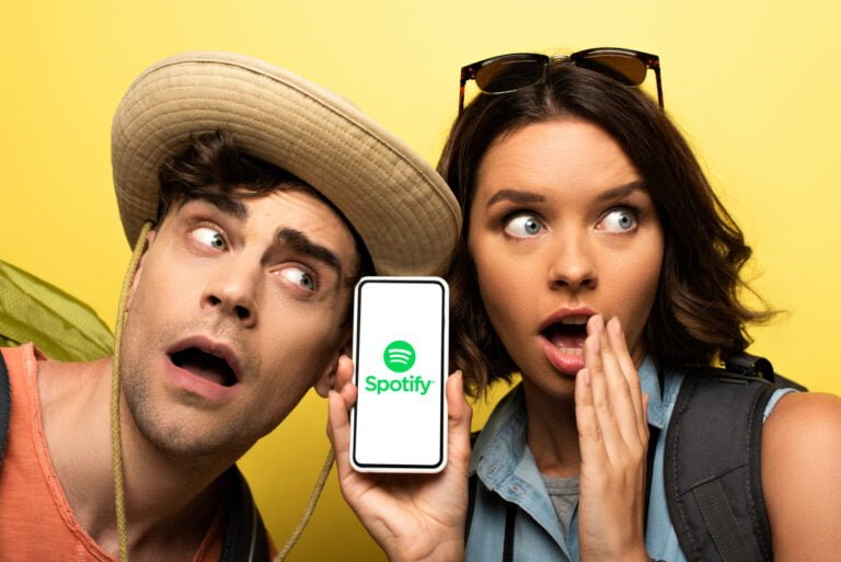 Kobieta i mężczyzna jasnej karnacji ze zdziwionymi minami, stykają się głowami na żółtym tle i patrzą w jedną stronę. Mężczyzna jest w słomkowym kapelusz, kobieta w ciemnych włosach ma na głowie okulary przeciwsłoneczne i trzyma smartfona, na którego ekranie jest zielone logo i Spotify Premium za darmo.