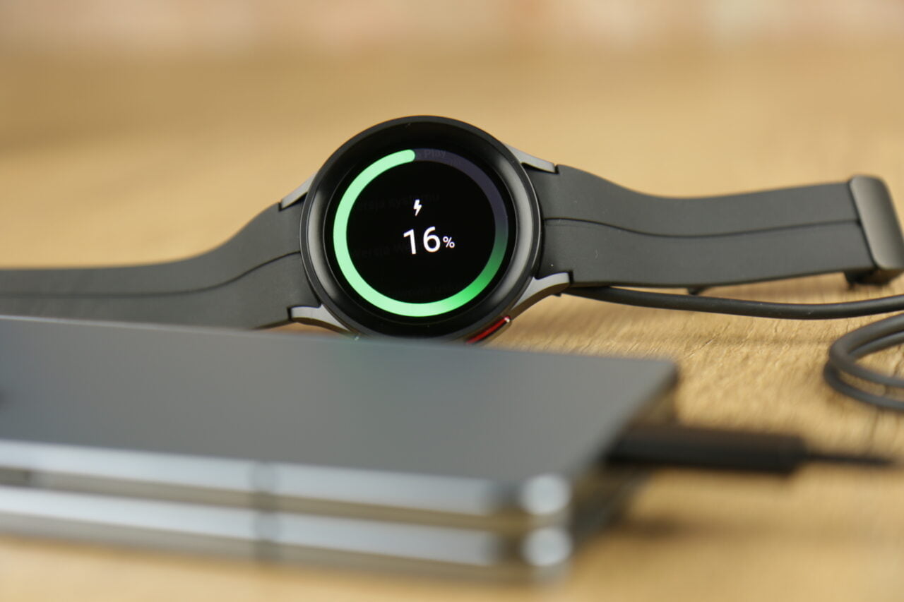 Czarny smartwatch leżący na szarym laptopie, z ekranem pokazującym 16% naładowania baterii.