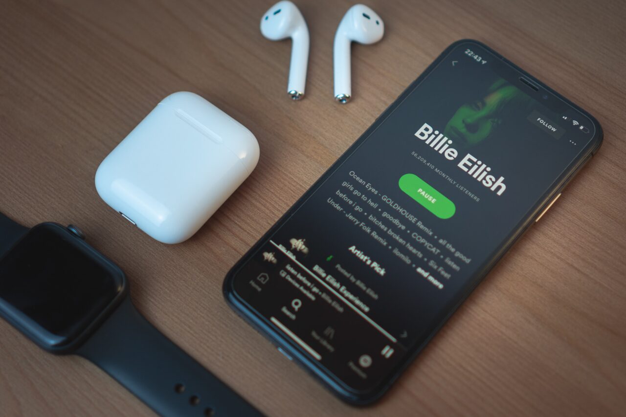 Smartfon leżący na drewnianym blacie wyświetlający profil artysty Billie Eilish na platformie streamingowej, obok słuchawki bezprzewodowe i opakowanie do ładowania oraz czarny smartwatch.