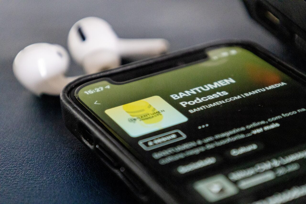 Smartfon wyświetlający ekran aplikacji podcastów o nazwie BANTUMEN, z białymi bezprzewodowymi słuchawkami umieszczonymi na czarnym tle obok telefonu.