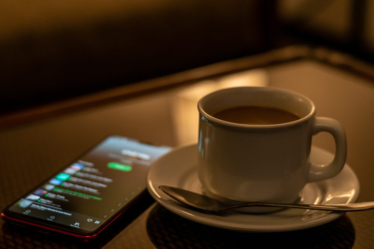 Filiżanka kawy na spodku z łyżeczką obok smartfona wyświetlającego listę utworów muzycznych.