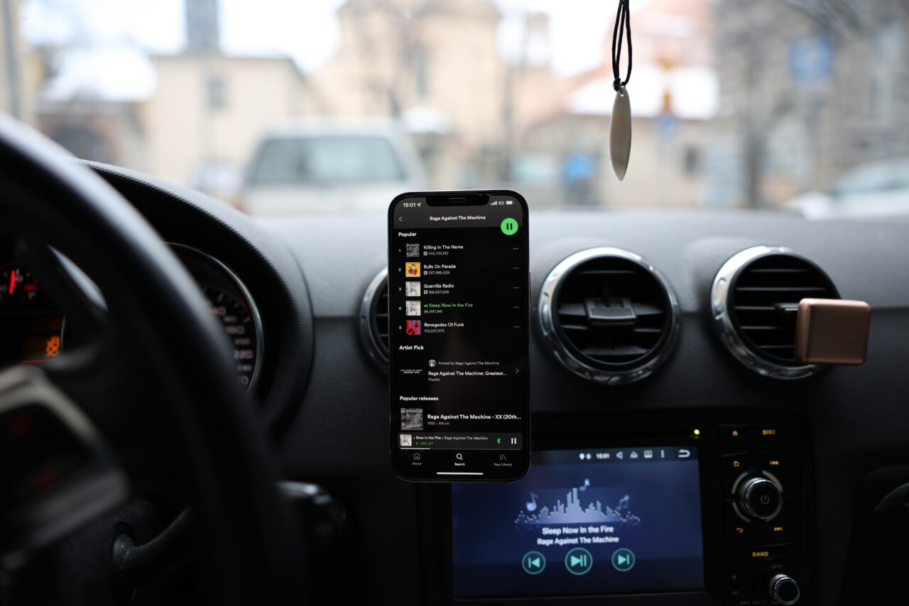 Wyświetlacz smartfona w samochodzie pokazujący aplikację Spotify z muzyką zespołu Rage Against The Machine, na pierwszym planie, z widokiem na deskę rozdzielczą i częściowo widoczną kierownicę w tle.