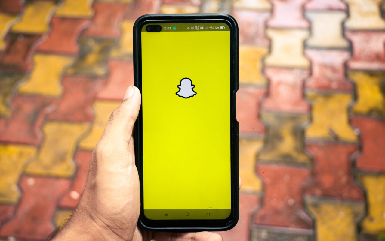 Osoba trzymająca smartfon z otwartą aplikacją Snapchat na ekranie startowym, widoczne logo Snapchat na żółtym tle, w tle rozmyte kolorowe płytki chodnikowe.