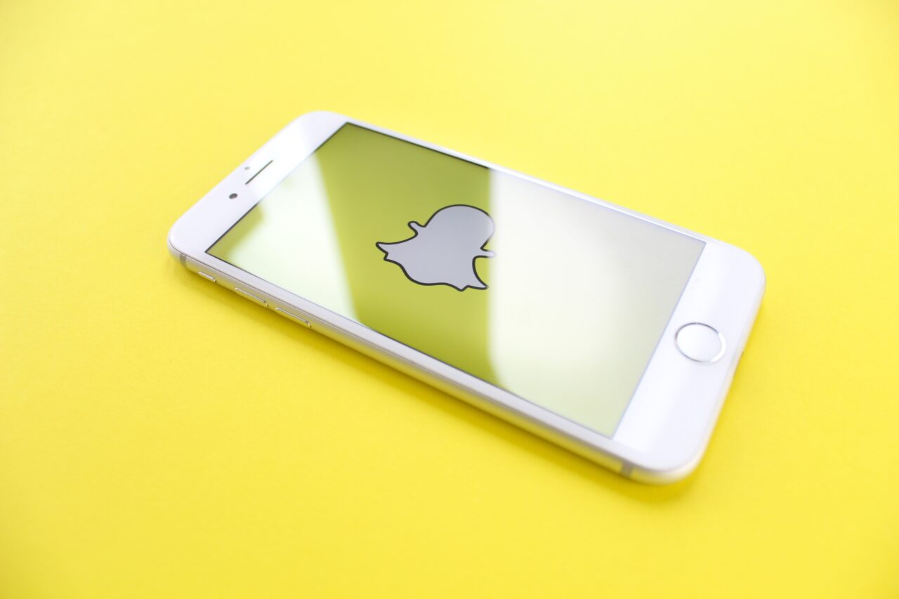 Biały smartfon na żółtym tle z otwartą aplikacją Snapchat na ekranie.