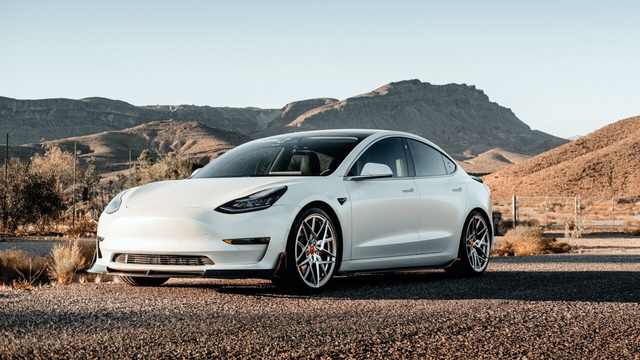 Biały samochód elektryczny marki Tesla zaparkowany na pustynnej drodze z górami w tle. Zdjęcie do tekstu opisujące autopilot Tesli i wypadki.