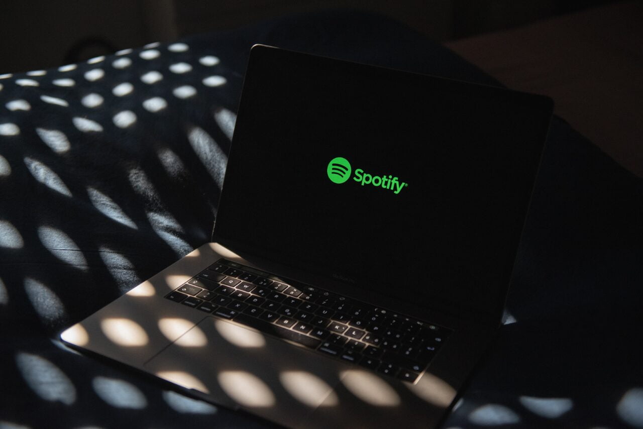 Laptop na łóżku z otwartą klapą wyświetlającą logo Spotify, oświetlony promieniami słonecznymi przechodzącymi przez roletę.