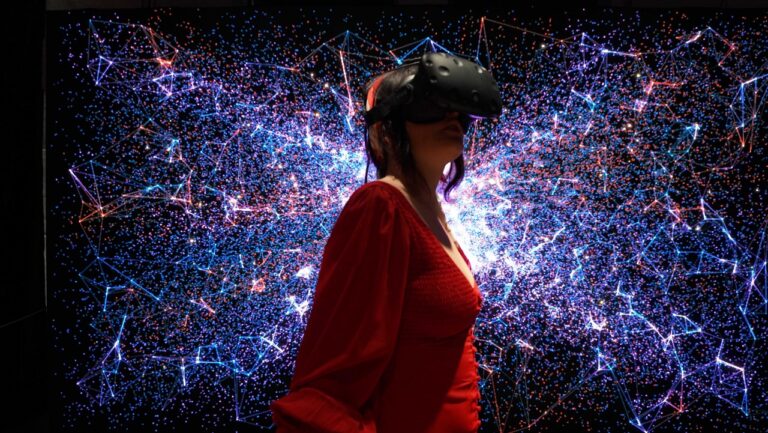 Kobieta w czerwonej sukience i z goglami VR na głowie stoi przed tłem z dynamicznymi wizualizacjami w postaci łączących się punktów i linii, imitujących sieć lub kosmos.