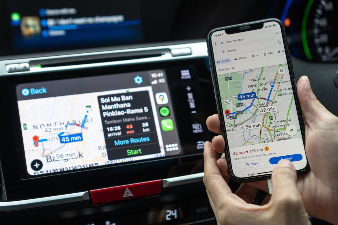 Dłoń trzymająca smartfon z włączoną aplikacją Google Maps wskazującą trasę z ruchem drogowym obok ekranu nawigacji samochodowej.