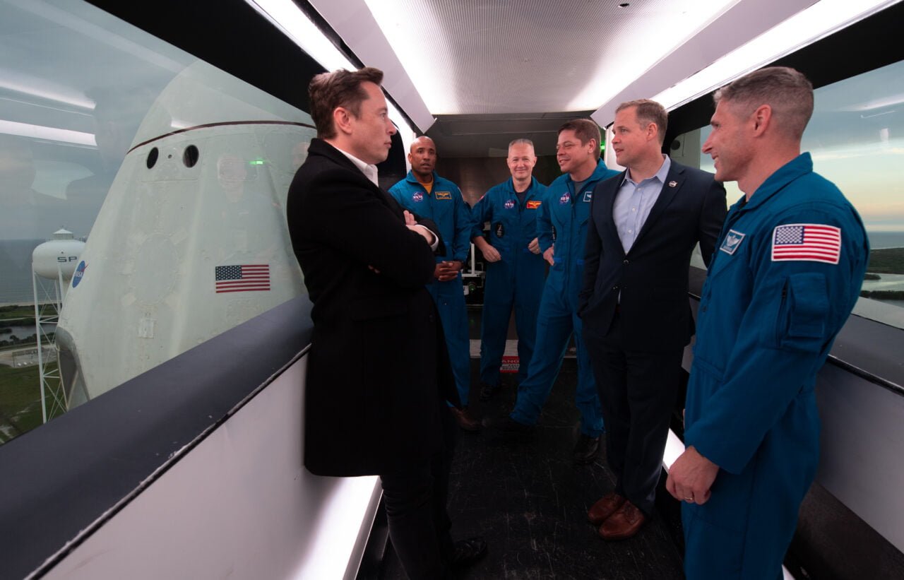 Elon Musk i astronauci. Grupa mężczyzn w garniturach i kombinezonach kosmicznych rozmawiająca w pomieszczeniu z widokiem na prom kosmiczny i wieżę startową.