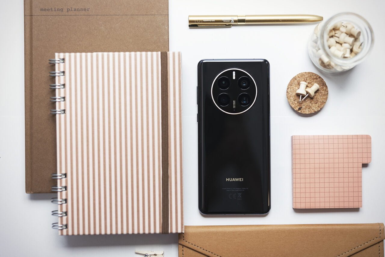 Zdjęcie do artykułu pt. jakiego smartfona kupić. Biurko z plannerem, czarnym smartfonem Huawei, złotym długopisem, słoikiem z zakrętkami, korkowym podkładem i różowym notatnikiem w kratkę.