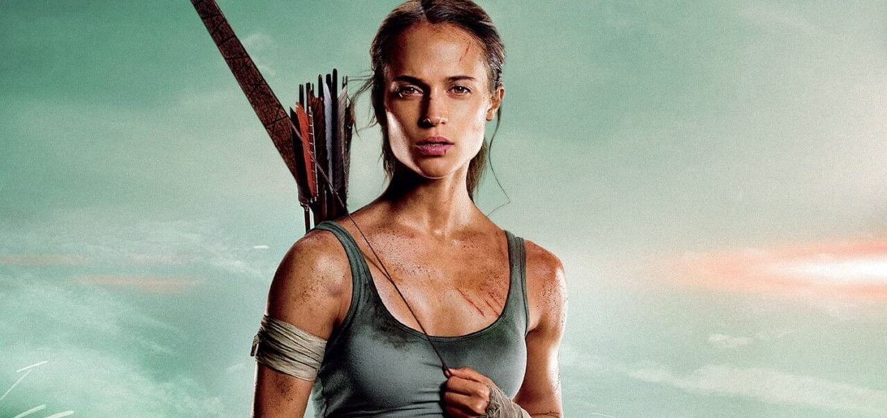 Alicja Vikander jako Lara Croft w filmie Tomb Raider z 2018 roku. Kobieta w zielonym topie z postrzępionym i owiniętym bandażem ramieniem, z kołczanem strzał na plecach na tle nieba.