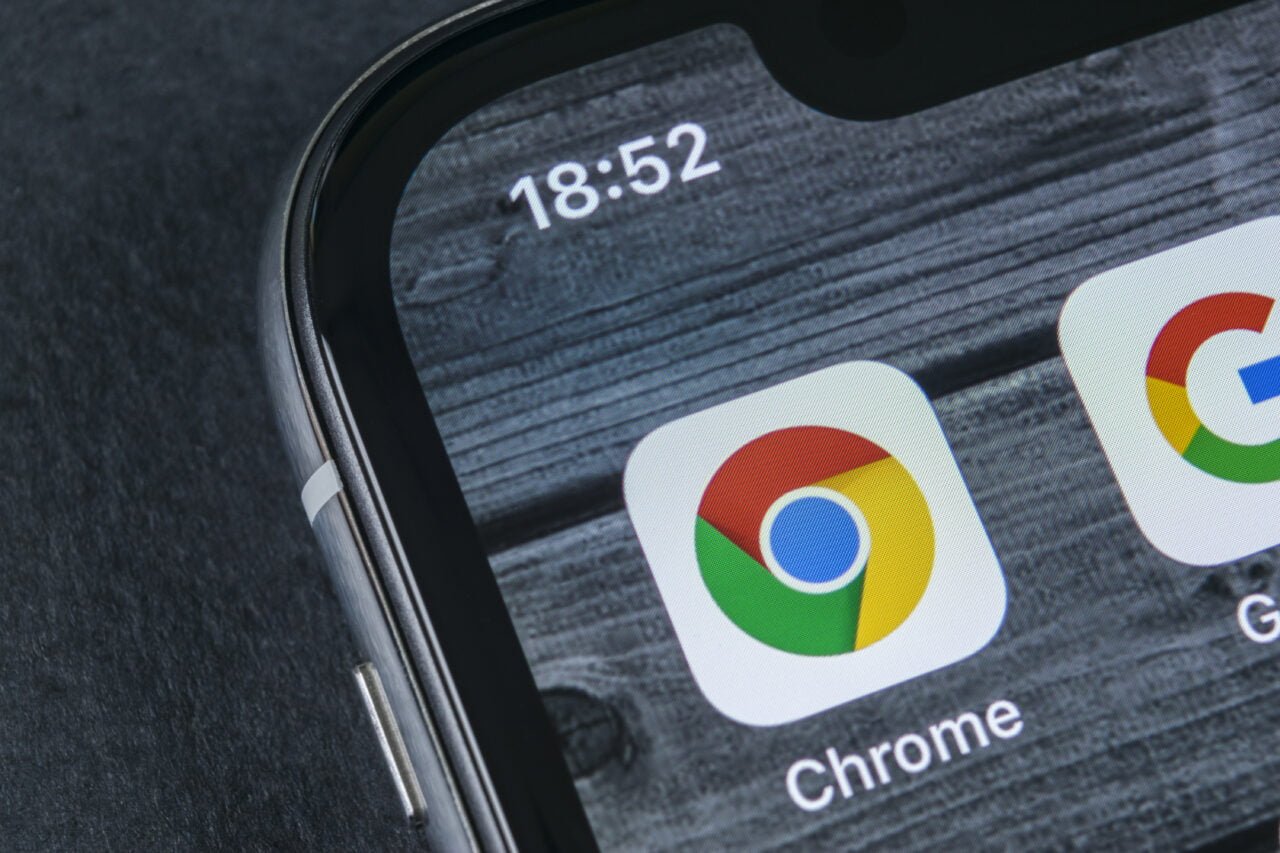 Parte da tela de um smartphone mostrando o ícone do navegador Google Chrome contra uma textura de madeira com o horário 18:52 visível na parte superior da tela.