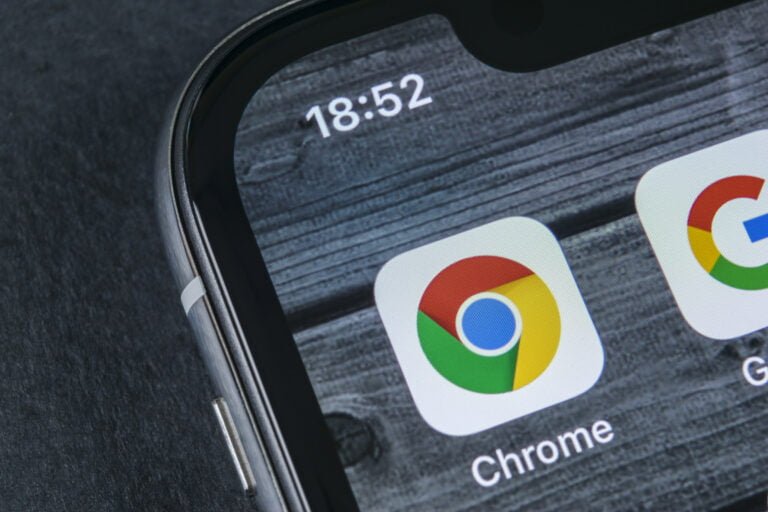 Część ekranu smartfona pokazująca ikonę przeglądarki Google Chrome na tle drewnianej tekstury z widocznym czasem 18:52 w górnej części ekranu.