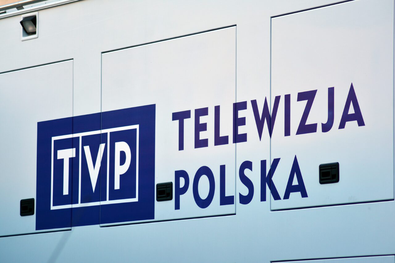 Biała ściana pojazdu transmisyjnego z niebieskimi napisami "TVP" i "TELEWIZJA POLSKA".