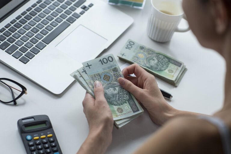 Osoba liczy banknoty o nominale 100 złotych na rachunki za prąd przy biurku, na którym znajduje się laptop, kalkulator i filiżanka.