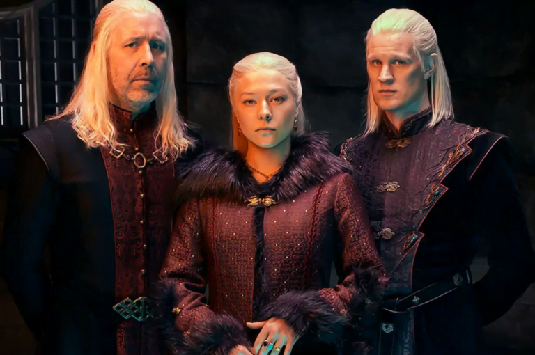 Trójka ludzi o bladych twarzach i blond włosach w historycznych, bogato zdobionych strojach, stoją ramię w ramię. Pochodzą z drugiego