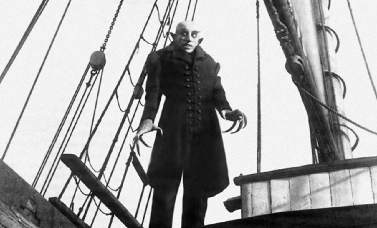 Czarno-biały obraz z filmu Nosferatu przedstawiający mężczyznę w charakterystycznym dla niemej ery filmu, gotyckim stylu stroju, stojącego na pokładzie statku z groźnym wyrazem twarzy i przerażającym makijażem przypominającym wampira.