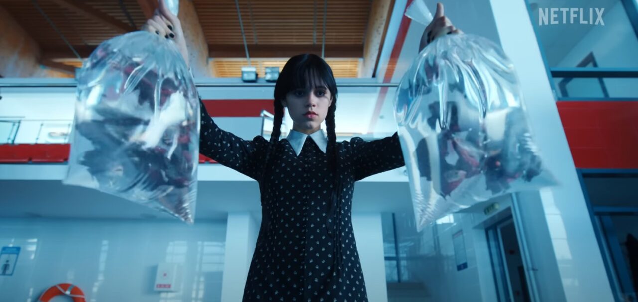 Wszyscy czekamy na drugi sezon Wednesday. Dziewczyna w ciemnej sukience z białym kołnierzykiem i warkoczami trzyma w wyciągniętych rękach dwie przezroczyste plastikowe torby z futurystycznymi maseczkami. Tło to współczesne wnętrze z elementami czerwieni i błękita, logo Netflix w prawym górnym rogu.