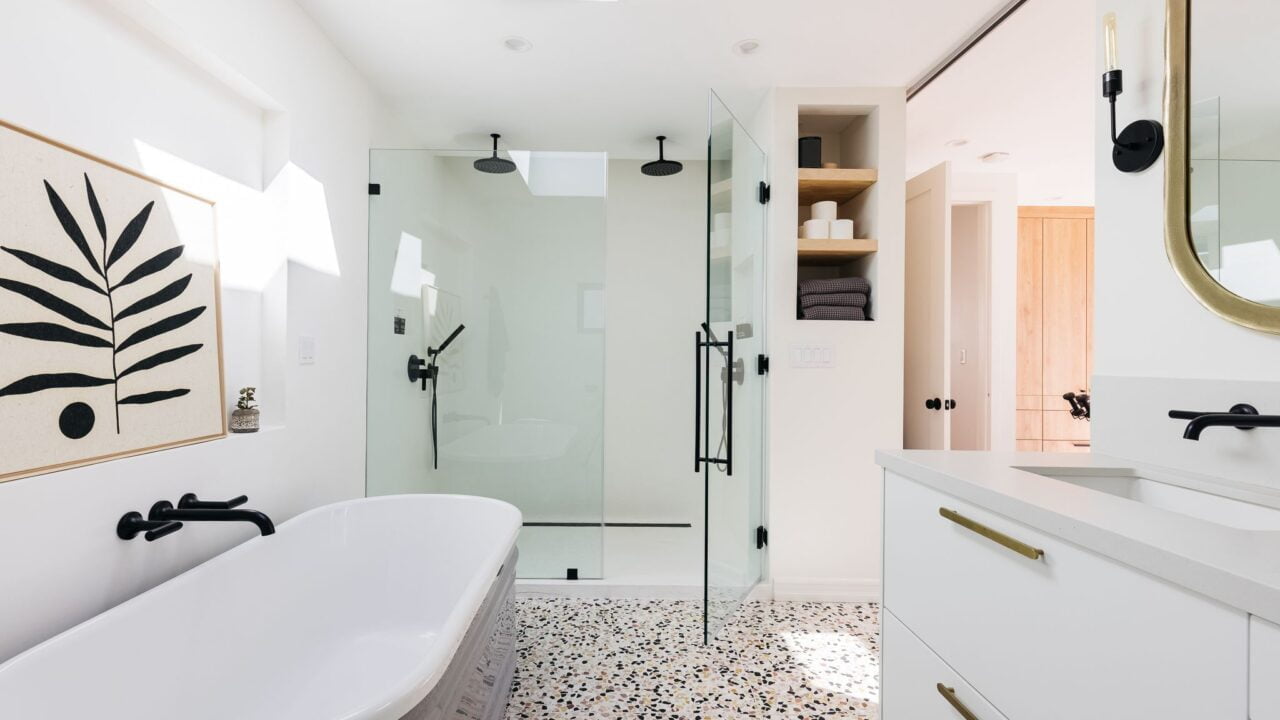 Nowoczesna łazienka z białymi ścianami, wolnostojącą wanną, prysznicem ze szklanymi drzwiami, podwójną umywalką na długiej szafce, okrągłym złotym lustrem oraz ozdobnym obrazem na ścianie.  Każde urządzenie sprawia, że rachunki za prąd są wyższe