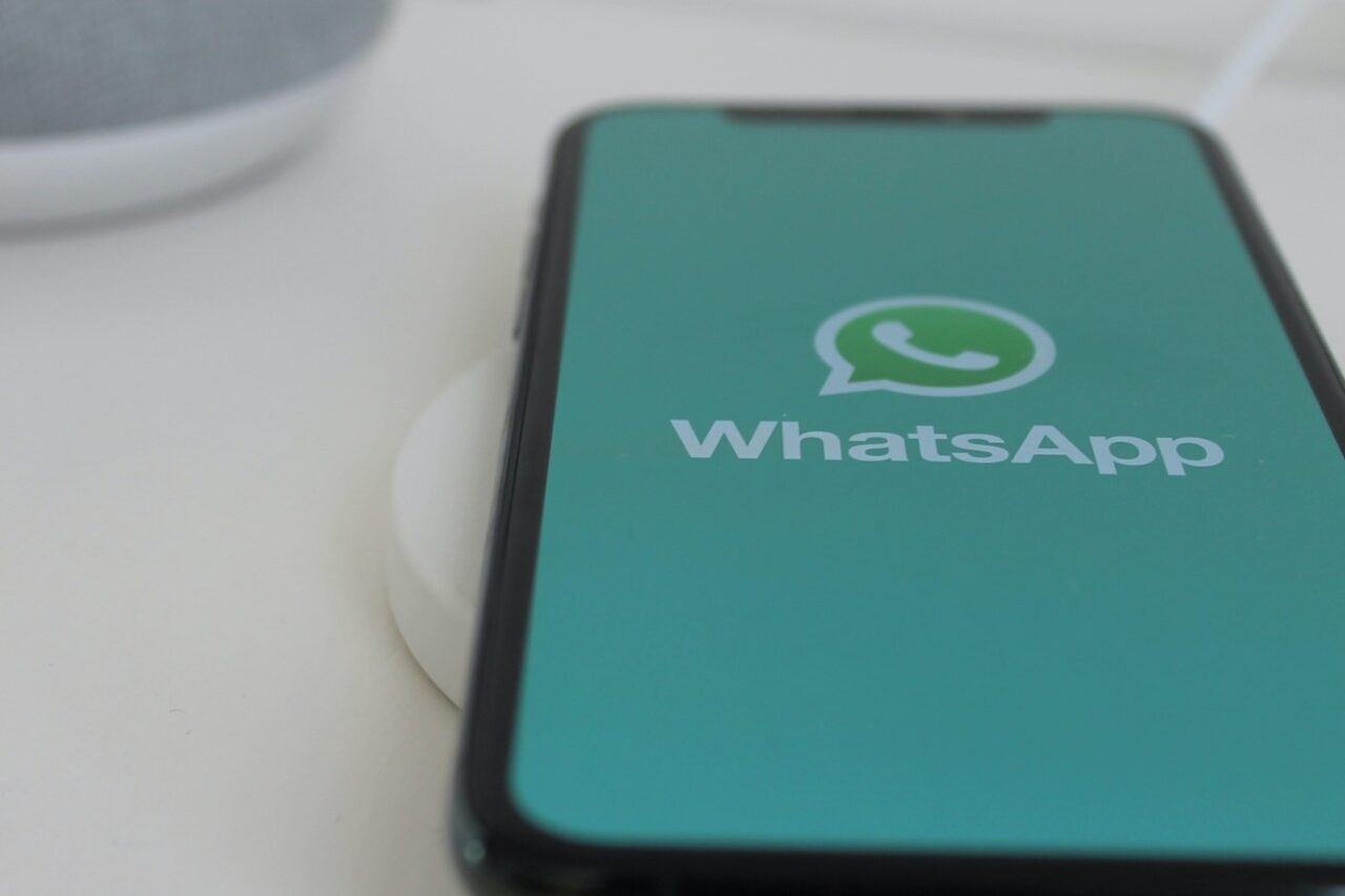 Smartfon na białym tle z otwartą aplikacją WhatsApp na ekranie, podłączony do białej ładowarki indukcyjnej, w tle częściowo widoczny głośnik inteligentny.