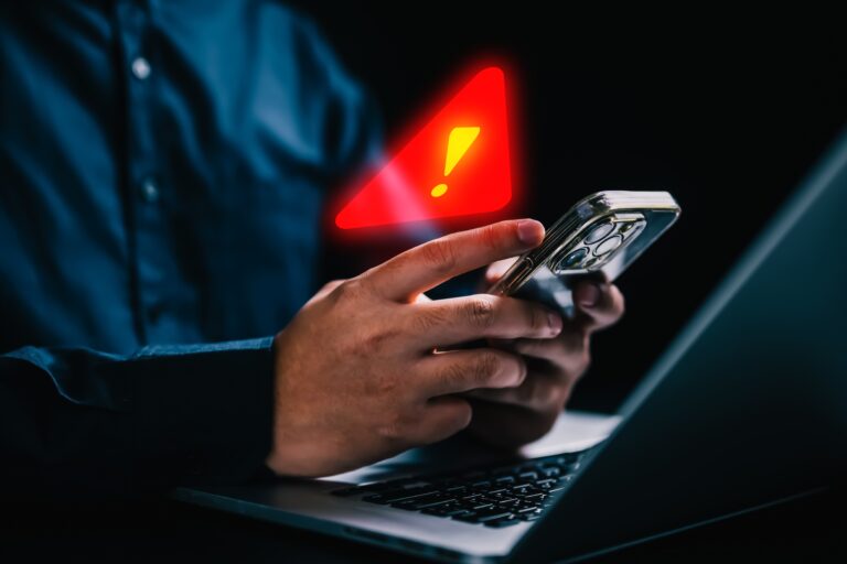 Osoba trzymająca smartfon, na ekranie którego wyświetla się czerwony trójkąt z wykrzyknikiem, w tle laptop.